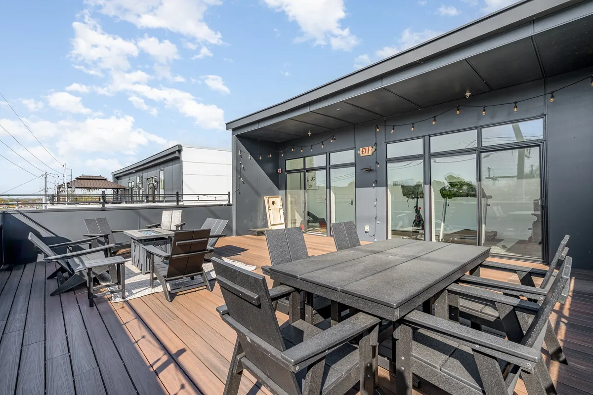 A stylish rooftop patio with outdoor dining table, reclining chairs, and string lights creating a cozy, welcoming ambience. The space is designed for social gatherings, relaxation, and evening events under the stars.