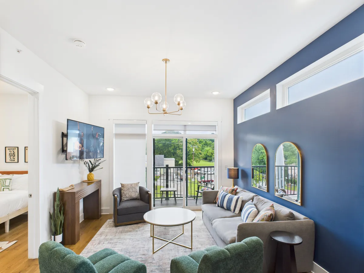 The image showcases a stylish, open-concept living space with a plush sofa adorned with decorative pillows, a statement chandelier overhead, expansive windows filling the room with natural light, a mirrored wall accent, and a sleek television. The atmosphere is cozy yet modern, perfect for both relaxation and social gatherings.