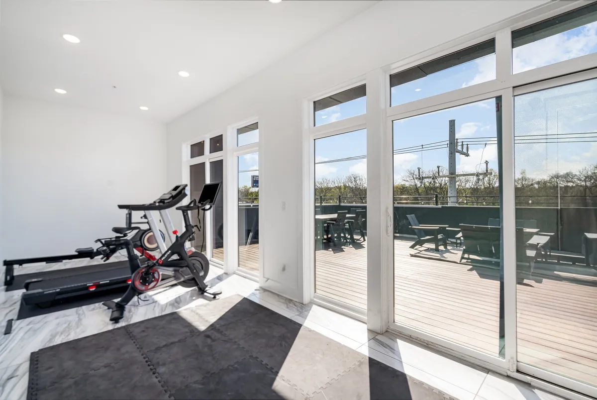 A modern gym space filled with exercise equipment, large windows that let in natural light, and a visible adjacent deck area. The atmosphere is bright and motivational, perfectly suited for maintaining an active lifestyle within the building.