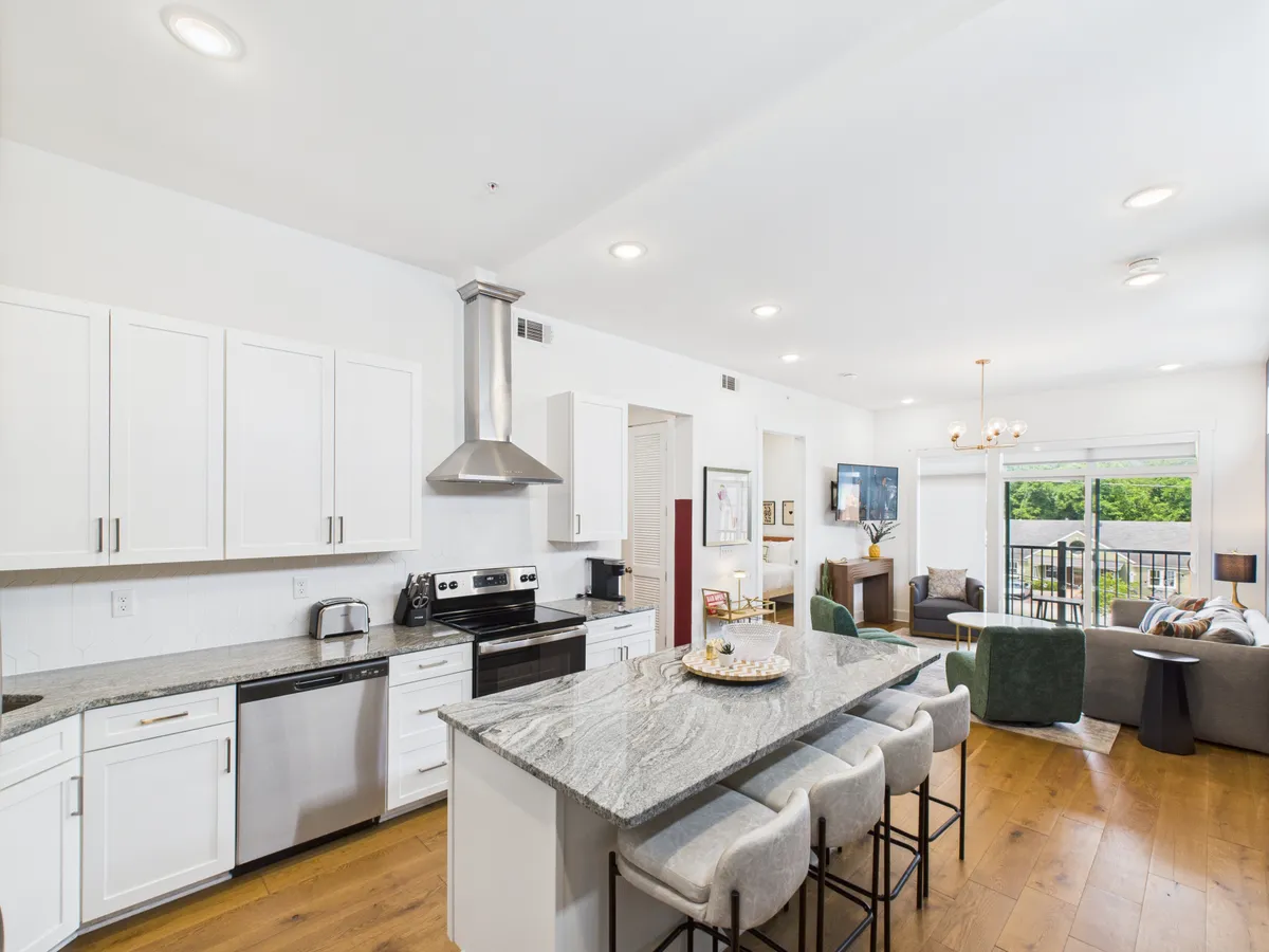 A bright, contemporary open-plan space showcasing a kitchen island with stainless steel appliances, large windows flooding the room with natural light, and a comfortable seating area that blends seamlessly with the cooking and dining zones. The area reflects both modernity and warmth, perfect for gatherings and shared meals.