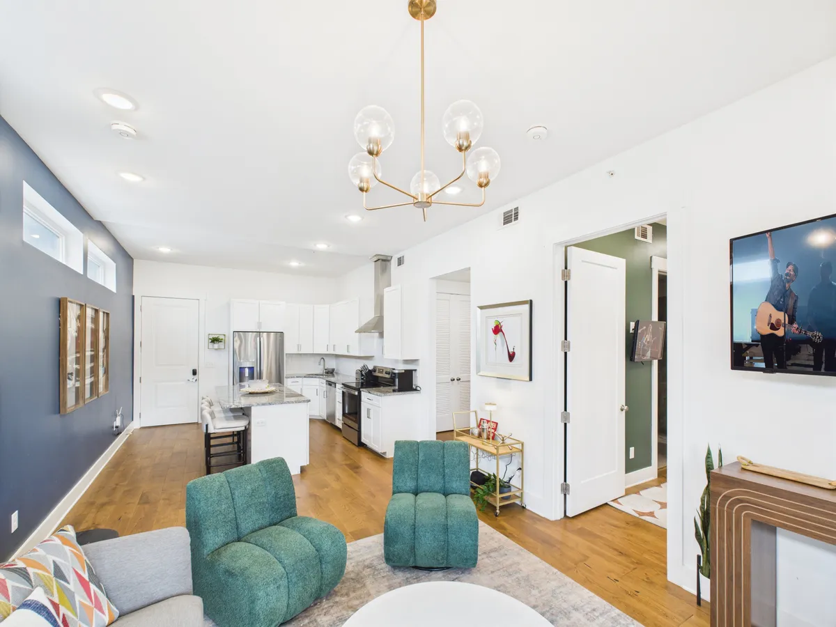 A cozy and stylish living area featuring green upholstered dining chairs, a contemporary chandelier, television, visible kitchen area, and modern wall art. The layout fosters easy movement and conversation between the living, dining, and kitchen spaces, reinforcing the open-concept feel.