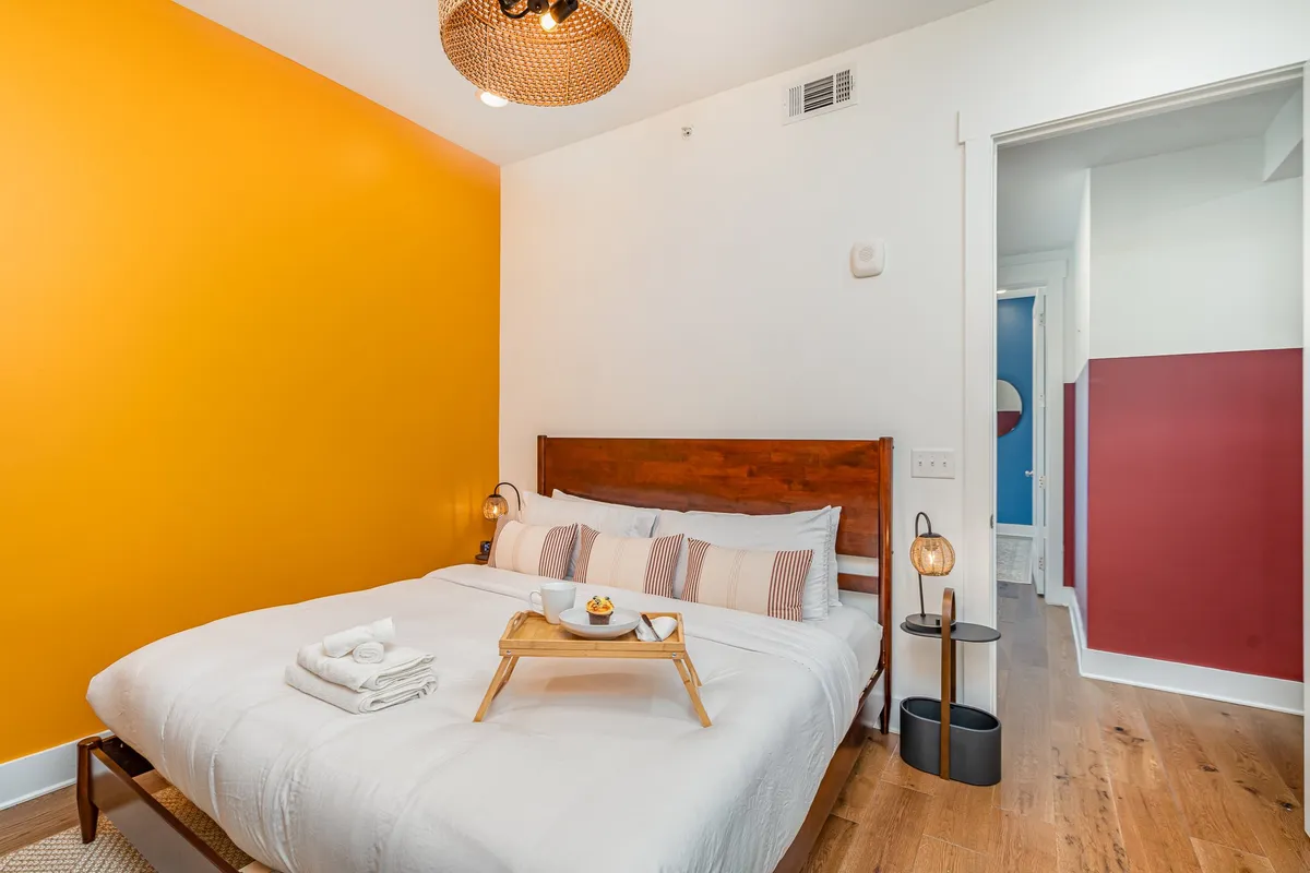 A vibrant bedroom featuring a bed with crisp white bedding, an orange accent wall, a stylish wooden frame, pendant lighting, and a breakfast tray, all bathed in natural morning light.