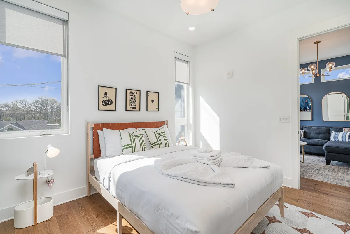 A tranquil bedroom scene with a plush bed, decorative wall art, a nightstand, and a large window allowing sunlight to softly illuminate the space. The neutral tones and cozy textures create a welcoming atmosphere ideal for rest and relaxation.